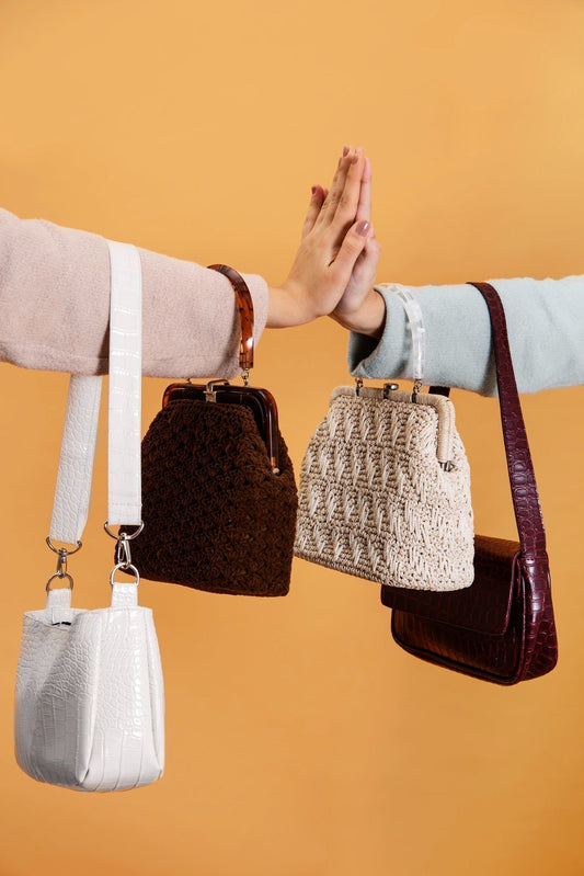 Two people high-five while displaying four luxury handbags - woven, crochet, and crocodile-textured on orange backdrop.