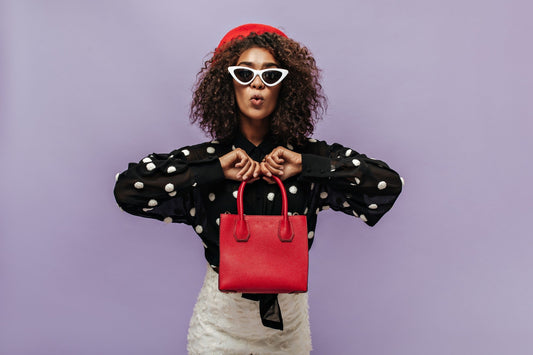 Stylish woman wearing a red beret, white cat-eye sunglasses, and a black polka dot blouse, holding a bright red handbag against a pastel purple background.