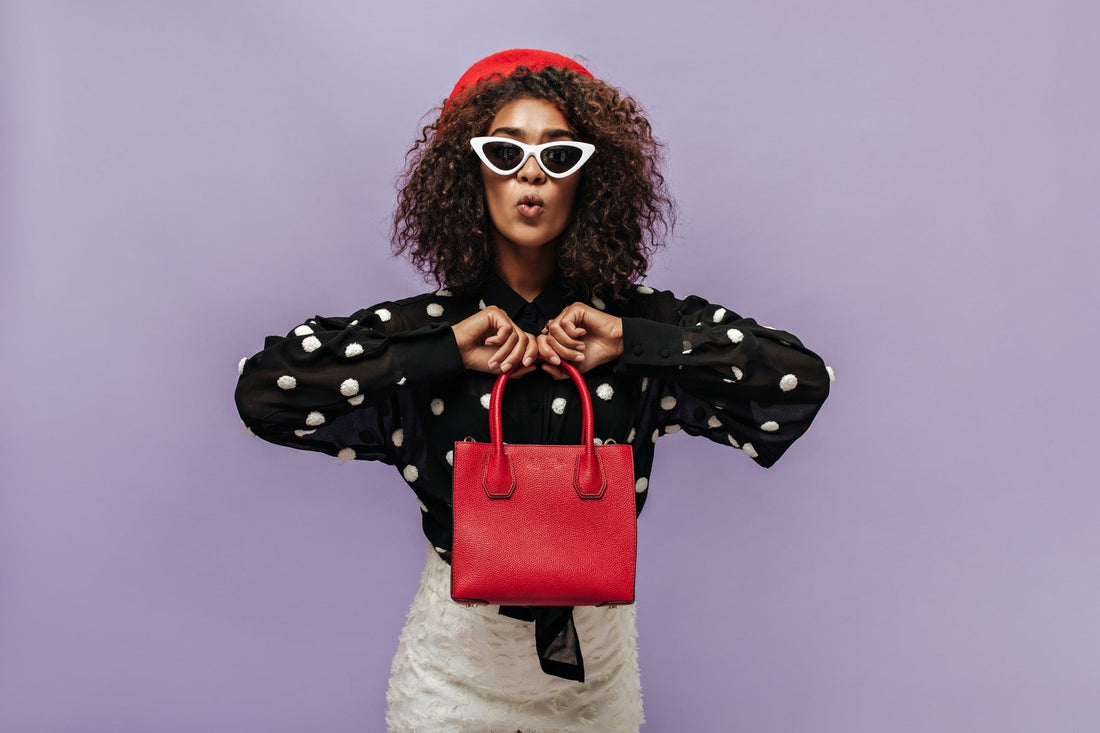 Stylish woman wearing a red beret, white cat-eye sunglasses, and a black polka dot blouse, holding a bright red handbag against a pastel purple background.